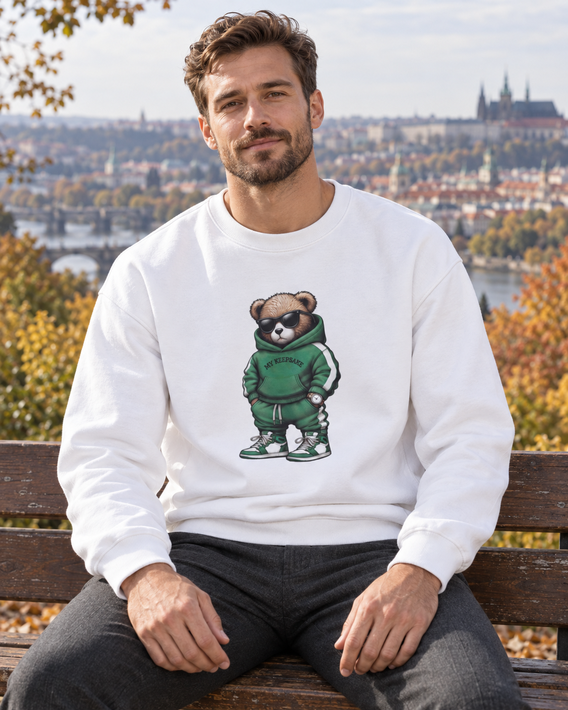Muž v bílé mikině s medvídkem My Keepsake na lavičce v pražském parku.